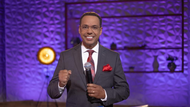 Stephen Colbert, Moderator von *The Daily Show with Trevor Noah*, steht auf einer Bühne und hält ein Mikrofon, trägt einen Anzug, Krawatte und Brille, während im Hintergrund ein Scheinwerfer und ein Ständer zu sehen sind.