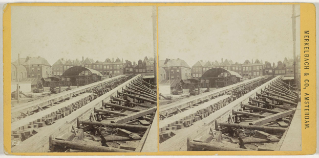 Schwarz-weißes Foto einer Zugschiene mit Gebäuden im Hintergrund, Text "Merkelbach & Co, Co. Amsterdam" auf der rechten Seite und verstreuten Holzobjekten im Vordergrund.