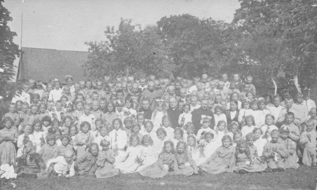 Schwarz-weiß-Fotografie einer Gruppe von Kindern, die vor einem Haus mit Bäumen und einem klaren Himmel posieren.