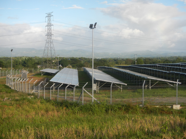Ein Solarpanel-Feld, das von einem Zaun umgeben ist, mit Gras und Pflanzen im Vordergrund, Bäumen und Übertragungsmasten mit Drähten im Hintergrund und einem bewölkten Himmel.