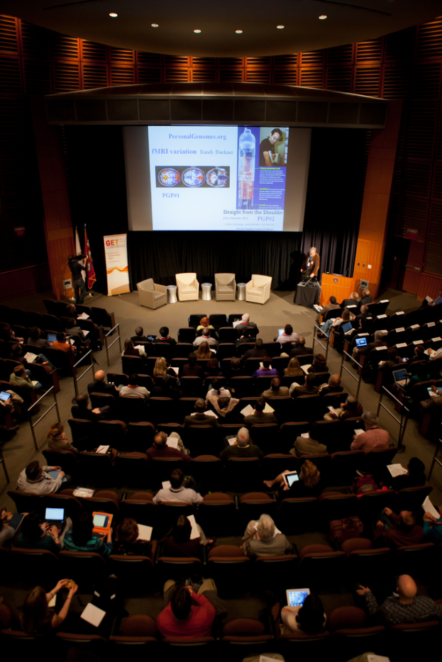 Ein mit sitzenden Menschen gefüllter Hörsaal, beleuchtet von vielen Lichtern, mit Fahnen links, einem Podium rechts und einer Werbetafel links.