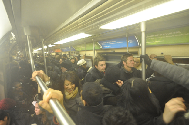Eine Gruppe von Menschen steht in einem Zug, hält sich an Metallstangen fest, mit Text auf Schildern, Fenstern, Dach, Lampen und anderen Gegenständen zu sehen.