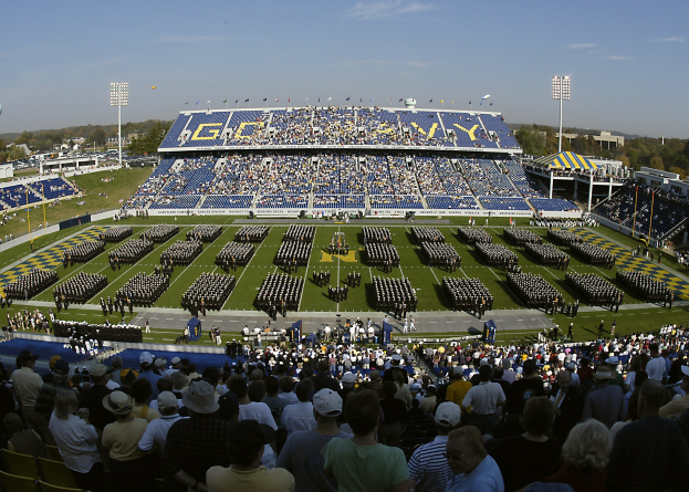 Große Menschenmenge bei einem Fußballspiel auf einem grünen Stadionrasen unter einem klaren blauen Himmel mit Flutlichtmasten, Bäumen und Gebäuden im Hintergrund.