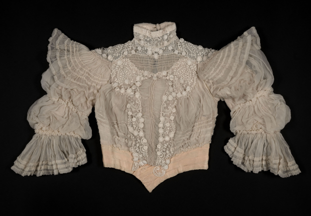 Eine weiße Bluse mit Rüschen-Ärmeln und Spitzenkragen, die einen zarten Spitzenbesatz am Kragen und den Ärmeln aufweist, vor einem schwarzen Hintergrund.