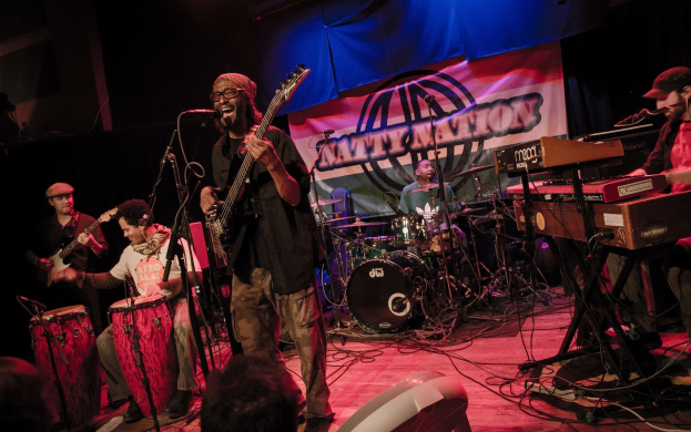 Eine Live-Band performt auf der Bühne mit Mikrofonen und Ständern, während das Publikum unten zuschaut; ein Banner hängt im Hintergrund.