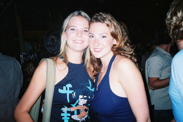 Zwei Frauen in blauen Kleidern, die lächeln, wobei eine ein Objekt trägt, mit ein paar anderen Menschen im dunklen Hintergrund.