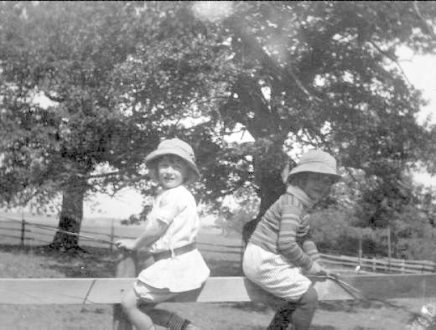 Zwei junge Jungen in Mützen und Baseball-Uniformen stehen in einem grasbewachsenen Park und halten Schläger, mit Bäumen und einem Zaun im Hintergrund, in Schwarz-Weiß.