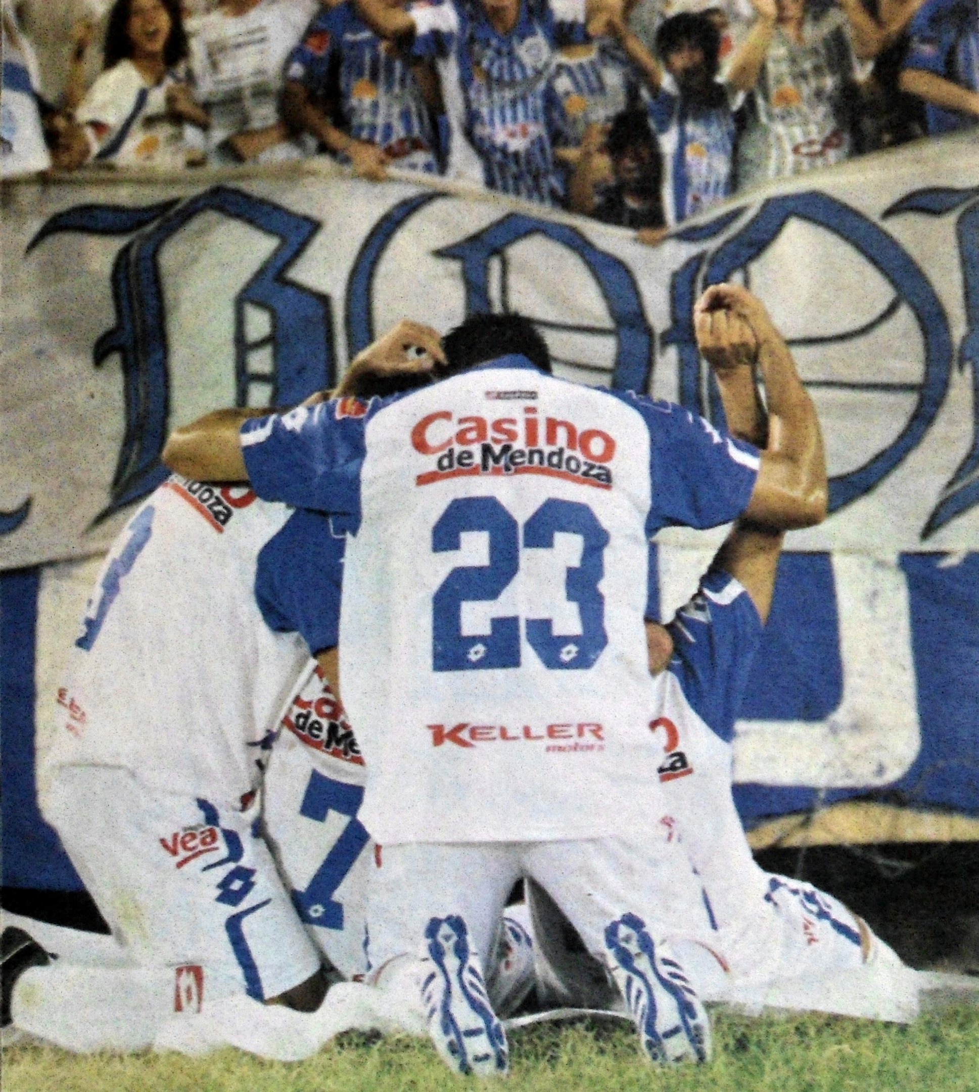 Gruppe von Fußballspielern, die auf dem Boden vor einer jubelnden Menge feiern, mit einem Banner mit Text im Hintergrund.