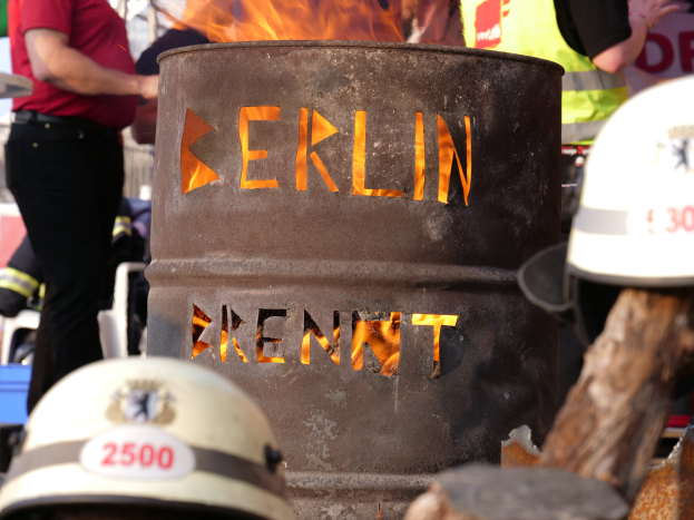 Eine Gruppe von Menschen um ein Feuerhydranten-Schild mit der Aufschrift "Berlin", mit einem brennenden Fass im Vordergrund, Helme unten und einem hölzernen Objekt auf der rechten Seite.