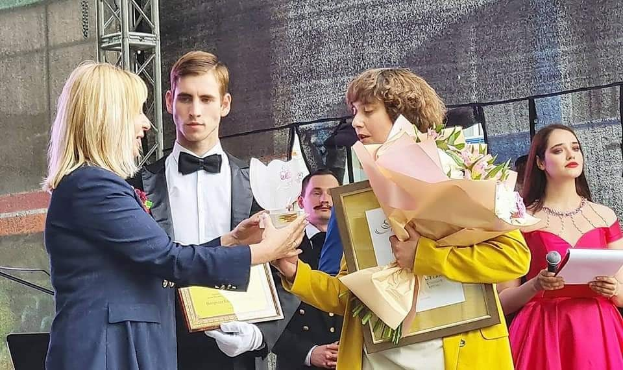 Gruppe von Menschen auf einer Bühne, eine in gelbem Kleid mit Blumen, eine andere mit Papieren und Mikrofon, eine Person links im blauen Blazer, mit Geländer und Wand im Hintergrund, wahrscheinlich bei einer Preisverleihung.