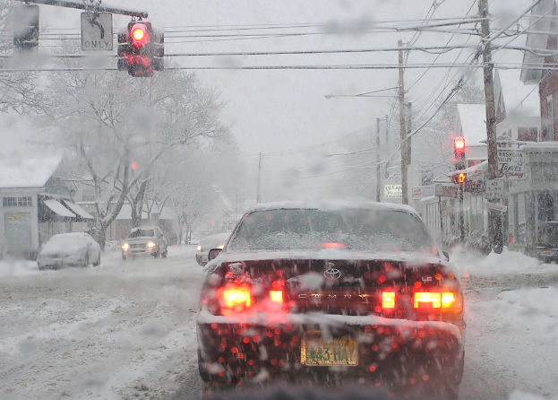 Eine schneebedeckte Straße mit spärlichem Fahrzeugverkehr, gesäumt von schneebedeckten Bäumen und Häusern auf beiden Seiten.