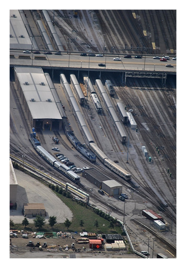 Eine Brücke mit ein paar Fahrzeugen darauf, Eisenbahnschienen mit ein paar Zügen darunter und eine Landschaft mit Gebäuden, Bäumen, Pfählen und verschiedenen Gegenständen und Fahrzeugen auf dem Boden.