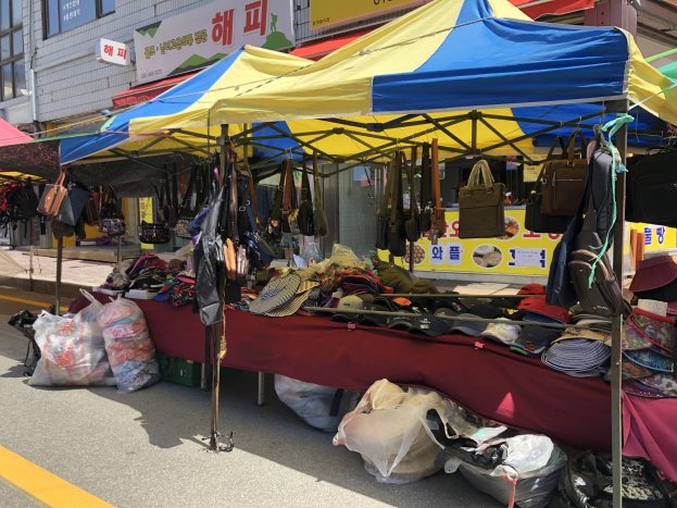 Ein Straßenmarkt mit Zelten, die Taschen, Hüte und andere Gegenstände präsentieren, vor einem Hintergrund von Gebäuden mit Fenstern und Texttafeln.