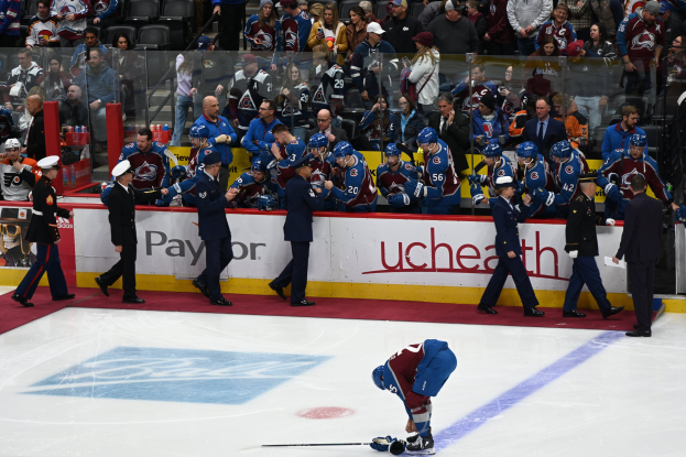 Eishockeyspieler auf dem Eis umgeben von Menschen mit Mützen und Stöcken, mit Zuschauern im Hintergrund und Brettern mit der Aufschrift "Colorado Avalanche vs San Jose Sharks - 3/24/15 NHL Pick, Odds, and Prediction".