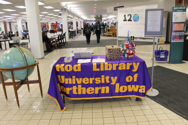 Eine Ausstellung mit einem Tisch, auf dem verschiedene Landesflaggen, eine Box, Papiere und ein Banner stehen, eine Kugel zur Linken, ein Anschlagbrett zur Rechten mit einigen Menschen in der Nähe und mehreren Tischen und Stühlen im Hintergrund.