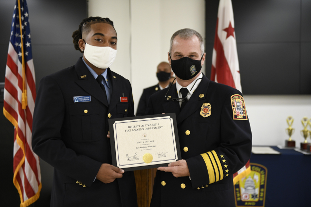 Zwei uniformierte Feuerwehrleute, einer identifiziert als Leiter der Feuerwehr des District of Columbia und der andere als stellvertretender Leiter, stehen Seite an Seite mit Masken und halten ein Zertifikat vor einem Tisch mit einer blauen Decke, Schilden und anderen Gegenständen, mit Flaggen und einer Wand im Hintergrund.