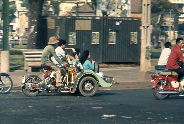 Mehrere Menschen auf Motorrädern auf einer Straße, mit einem Gehweg, einem Pfahl, einer Bank und einem Baumstamm dahinter und Gebäuden, einer Hütte und Gras im Hintergrund.