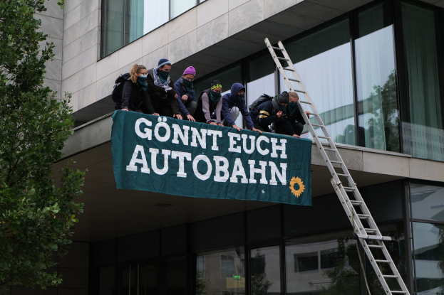 Gruppe von Menschen auf einem Gebäudedach mit einem Banner, mit einem Baum links und Glasfenstern im Hintergrund, wahrscheinlich protestierend im Support der deutschen Regierung.