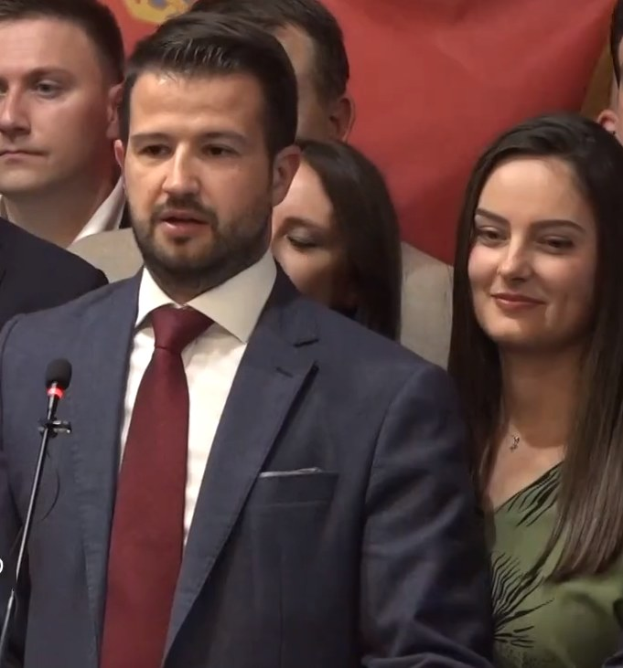 Ein Mann in Anzug und Krawatte spricht an einem Rednerpult mit einem Mikrofon zu einer Menge hinter einem Banner, wahrscheinlich über ein Regierungsverbot von Social Media.