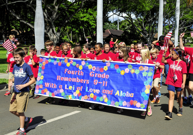 Gruppe von Kindern in roten Hemden, die eine Straße entlanggehen und eine Fahne sowie Flaggen tragen, mit Bäumen, Pfählen und Häusern im Hintergrund.