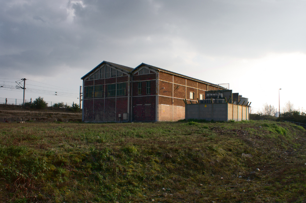 Eine Szene mit Gartenhäusern, Transformatoren, Strommasten mit Kabeln, Zäunen, einer Straßenlaterne, Bäumen und einem bewölkten Himmel.