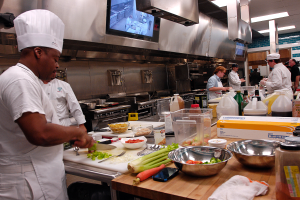 Eine Gruppe von Köchen in einer Küche beim Zubereiten von Essen, mit einem Koch im Vordergrund in einer weißen Schürze und Mütze, der ein Messer hält und von verschiedenen Utensilien, Schüsseln, Gemüse, einem Handy, Flaschen und anderen Gegenständen umgeben ist, mit einem Fernseher und Deckenlampen im Hintergrund.