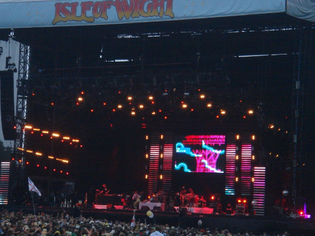 Eine Bühne mit Scheinwerfern, einem Bildschirm und Musikern, die Instrumente spielen, mit einem Banner oben, einer Menge unten und Fahnen mit Stangen und Luftballons drumherum.