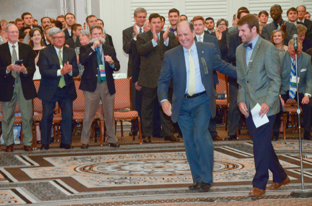 Zwei Männer im Anzug stehen mit einem Mikrofon rechts im Vordergrund, während im Hintergrund Menschen applaudieren und Stühle, Fenster und eine Tür zu sehen sind.