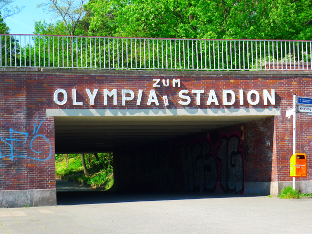 Der Eingang zum Olympiastadion in Berlin, Deutschland, mit einer Brücke mit Text, einem Metallzaun, einer Tafel, einer Box, Pflanzen, Gras, einer Baumgruppe und einem bewölkten Himmel.