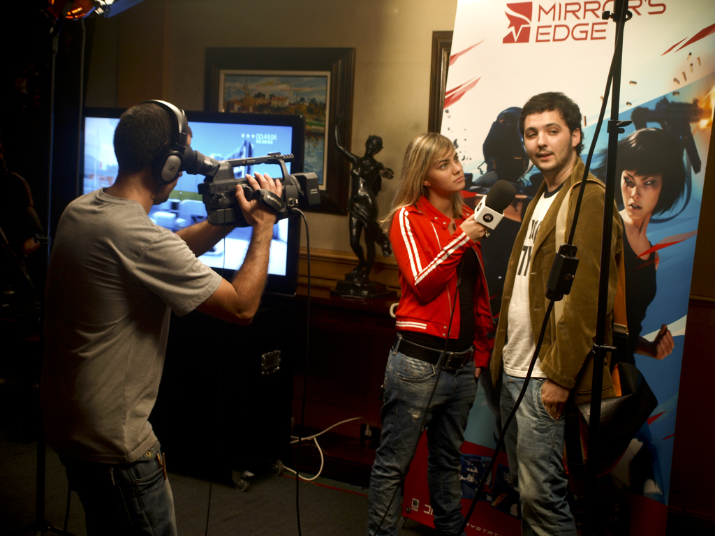 Ein Mann mit einer Videokamera und einem Headset filmt ein Interview zwischen einem Mann und einer Frau, wobei im Hintergrund ein Fernseher und ein Foto Rahmen zu sehen sind.