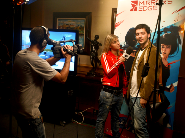 Ein Mann mit einer Videokamera und einem Headset filmt ein Interview zwischen einem Mann und einer Frau, wobei im Hintergrund ein Fernseher und ein Foto Rahmen zu sehen sind.