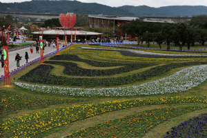 Ein großer Blumen Garten in einem Park mit Menschen, die spazieren gehen, umgeben von grünem Gras, Bäumen, Gebäuden, einer Brücke, Bergen und einem klaren blauen Himmel, mit Pfählen und Lichtern auf der linken Seite.