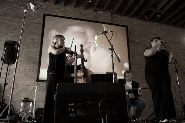 Eine Gruppe von Musikern in Schwarz, mit einem Mädchen, das Geige spielt, zwei Männern, die Gitarre spielen, im Hintergrund, und einer Frau, die rechts singt.