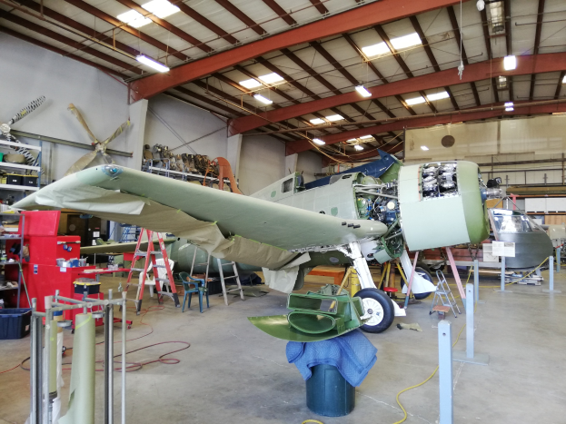 Ein großer Hangar mit verschiedenen Flugzeugen und Wartungsausrüstung, darunter eine Leiter, ein Tisch, ein Ventilator und Deckenlampen.