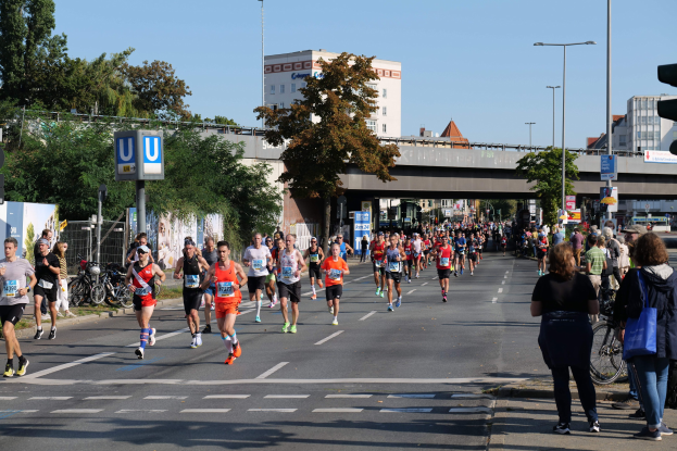 Gruppe von Menschen, die bei einem Marathon auf einer Straße mit Bäumen, Straßenmöbeln und Gebäuden unter einem klaren blauen Himmel laufen.