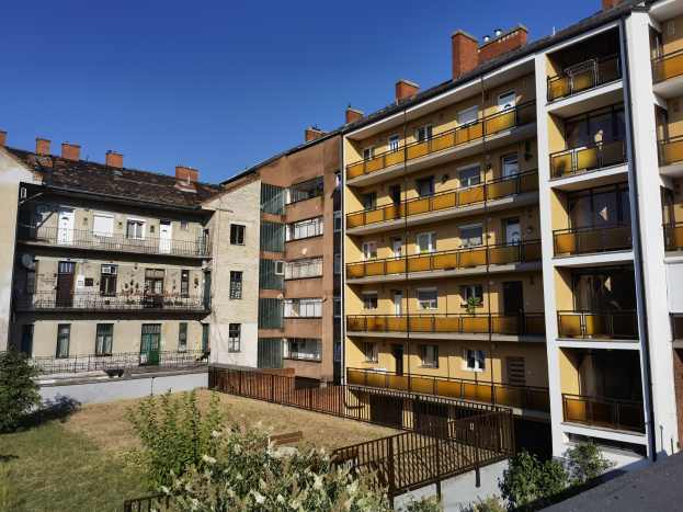 Ein Wohnhaus mit einem Innenhof, umgeben von saftigem Gras, Pflanzen und Bäumen, mit Fenstern, Türen und Geländern, und dem Himmel im Hintergrund.