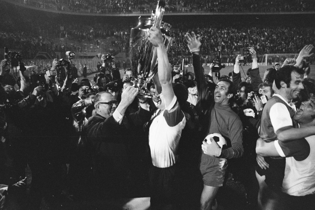 Schwarzes und weißes Bild einer Gruppe von Männern, die mit einem Pokal feiern, der zentrale Figur hält den Becher, während andere ihn umarmen, vor einem jubelnden Stadionpublikum mit Menschen, die Fotos machen.