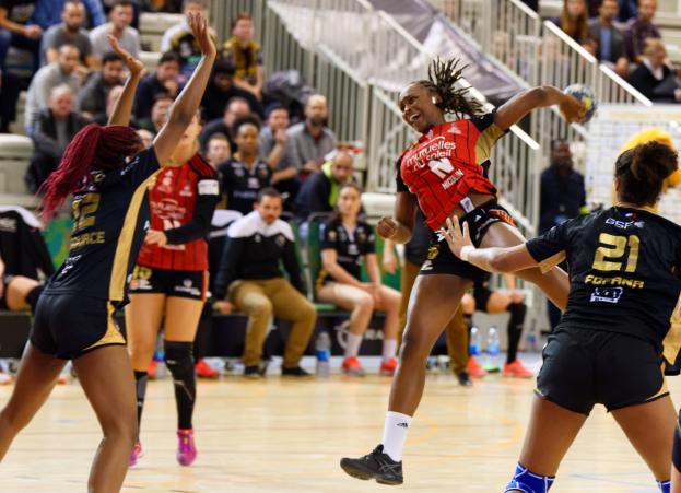 Eine Gruppe von Frauen, die Volleyball auf einem Feld mit einem Ball in der Mitte spielen, während Zuschauer auf Stühlen und Geländern im Hintergrund sitzen und Flaschen auf dem Boden liegen.