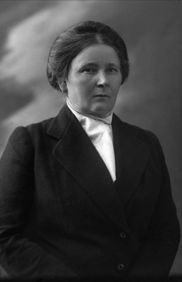 Schwarz-weiß-Foto einer Frau in einem Anzug, die mit entschlossener Miene und gefalteten Händen sowie einem Dutt steht und Suffragettenstärke verkörpert.