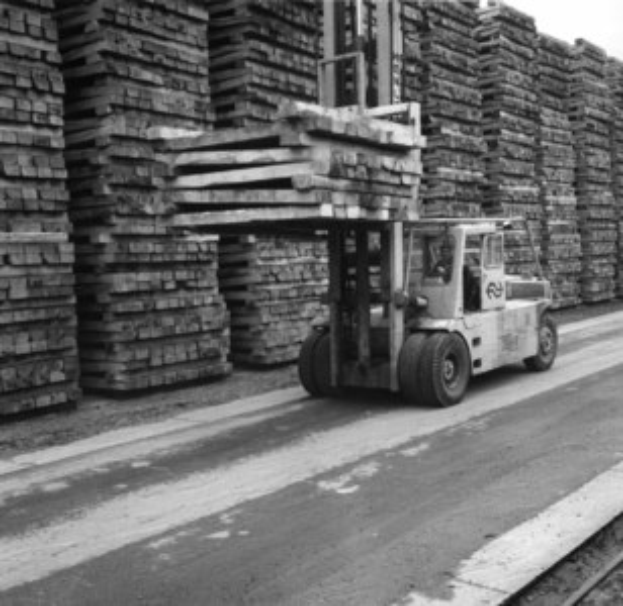 Schwarzes und weißes Foto eines Gabelstaplers mit einer Person darin, die eine Straße entlangfährt, neben einer Eisenbahnschiene, mit gestapelten Holzlatten im Hintergrund.