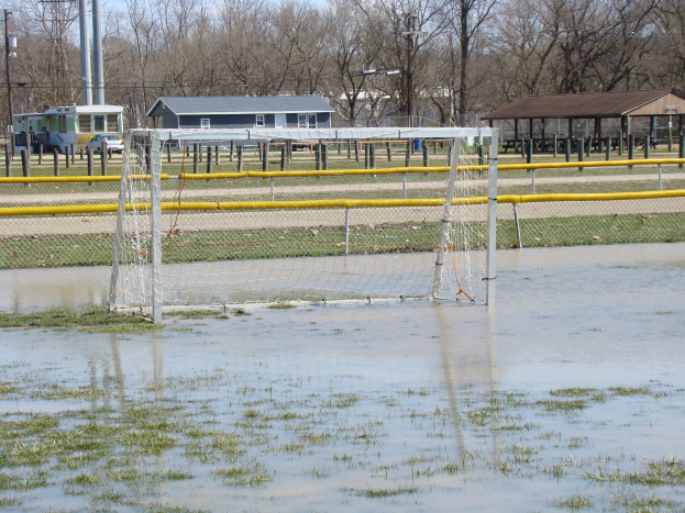 Ein Fußballtor steht in der Mitte eines überfluteten Feldes, umgeben von Gras und Wasser, mit Schuppen, Pfählen, Bäumen und Fahrzeugen im Hintergrund und einem klaren blauen Himmel.