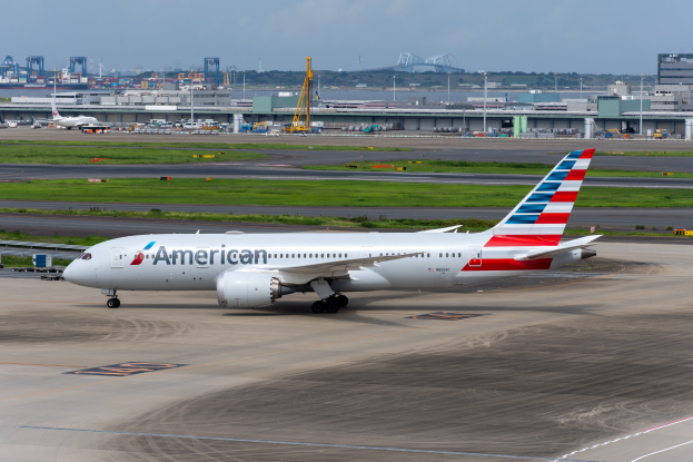 Ein American-Airlines-Flugzeug steht auf der Rollbahn eines Flughafens, umgeben von Gras, Fahrzeugen, Gebäuden, Masten, Kränen und anderen Gegenständen, mit Bäumen und einem klaren blauen Himmel im Hintergrund.