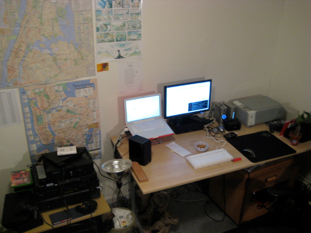 Ein Raum mit einem Tisch, auf dem ein Laptop, Lautsprecher, Monitor, Tastatur, Ball, Maus, Scanner und Papiere sowie ein weiterer Tisch mit Elektronikgeräten im Hintergrund stehen, mit Charts und Postern an der Wand.