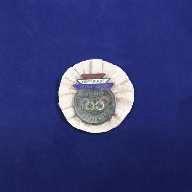 Blauer Stoff mit einem zentralen Olympischen Medaillenabzeichen, das das Logo und den Text enthält.