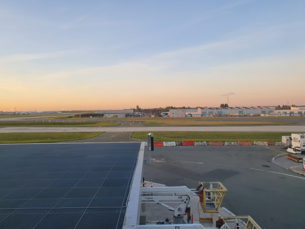 Luftbild eines Flughafens mit Fahrzeugen auf Straßen, Rasenflächen, Gebäuden, Bäumen und einem bewölkten Himmel, das eine Solarpaneel auf dem Dach des Gebäudes zeigt.
