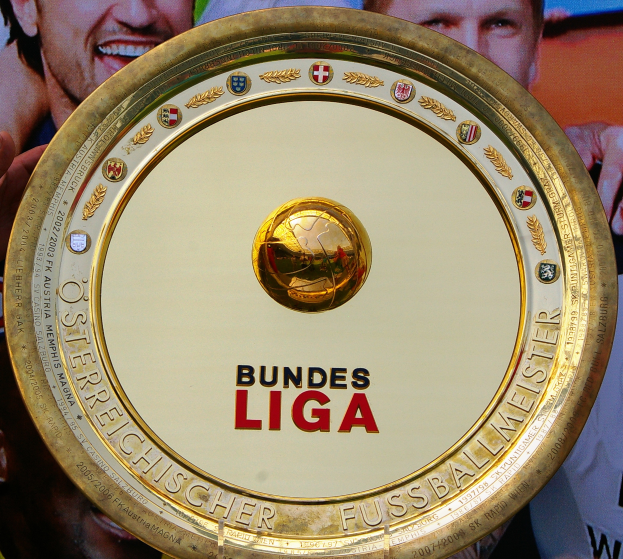 Ein Mann hält ein goldenes Schild mit der Aufschrift "Bundesliga" hoch, im Hintergrund ist ein Banner mit drei Personen zu sehen.