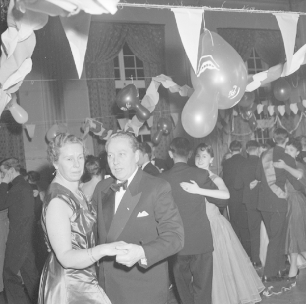 Ein Mann in einem schwarzen Blazer, weißem Hemd und schwarzen Hosen tanzt mit einer Frau auf einer Party, umgeben von Menschen, Ballons, Vorhängen und einem Fenster, auf einem Schwarz-Weiß-Bild.