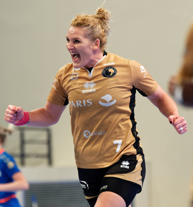 Eine Frau in braunem T-Shirt und schwarzen Shorts rennt auf einem Futsalplatz, feiert mit triumphierendem Gesichtsausdruck, während zwei andere Frauen im Hintergrund zu sehen sind.
