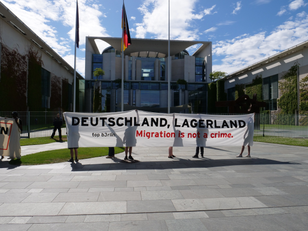 Gruppe von Menschen mit einem Banner mit der Aufschrift "Deutschland, Lagerland Migration ist kein Verbrechen" vor einem Gebäude mit Fenstern, umgeben von Gras, einem Metallzaun, Pflanzen, Bäumen und Fahnenmasten mit Flaggen, unter einem bewölkten Himmel.