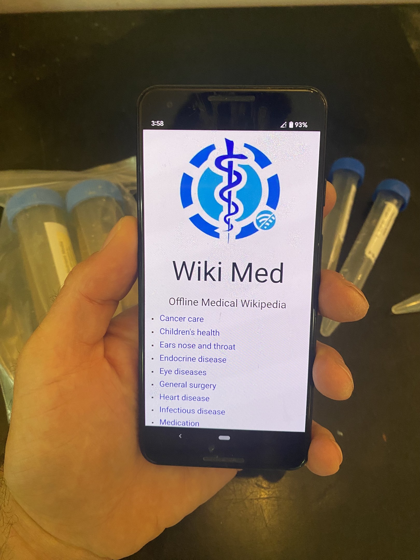 Eine Person mit einem Smartphone, auf dem die WikiMed-App geöffnet ist, mit Reagenzgläsern und anderen Gegenständen auf dem Tisch davor.